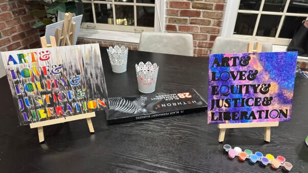 Two artworks on easels, featuring the text "ART & LOVE & EQUITY & JUSTICE & LIBERATION," with a book and paint palette nearby.