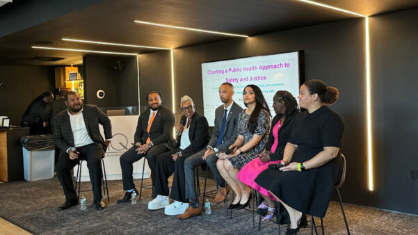 A panel discussion titled "Charting a Public Health Approach to Safety and Justice." Seven speakers, representing diverse backgrounds, sit on high stools in a contemporary venue. The backdrop features a presentation screen with the session title. The speakers engage with the audience, discussing public health and justice concepts.