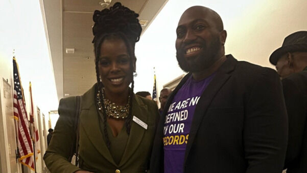 Two individuals are standing side by side in an indoor hallway. The woman on the left is wearing a fitted olive green suit and a statement necklace, smiling and holding a patterned handbag. The man on the right is also smiling and wearing a dark blazer over a purple t-shirt with the message "We are not defined by our records". They appear to be at a gathering, with U.S. flags visible in the background.