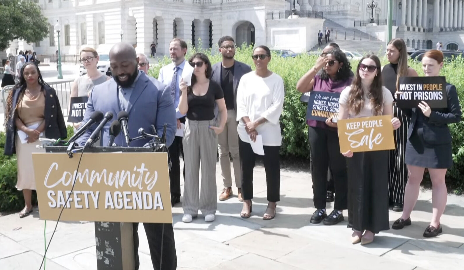 Dr. Kevin Beckford, Senior Associate of Partnerships and Engagement at the Pretrial Justice Institute, speaks at a press conference launching the Community Safety Working Agenda. Dr. Beckford is a Black man wearing a gray collared shirt, gray-blue suit jacket, and black pants. He is standing at a podium labeled “Community Safety Agenda.” Behind him, a diverse group holds signs that read “Keep People Safe” and “Invest in People, Not Prisons.”