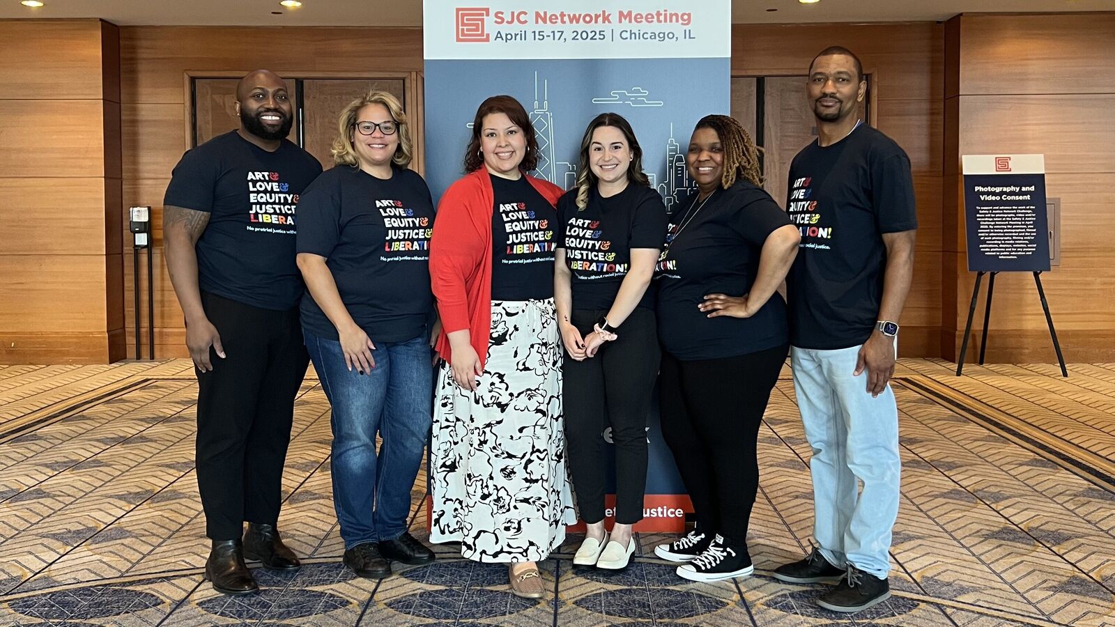 Six members of the PJI team smile in front of a large sign for the 2025 Safety and Justice Challenge Network Meeting. They all sport dark blue t-shirts with the message Art and Love and Equity and Justice and Liberation.